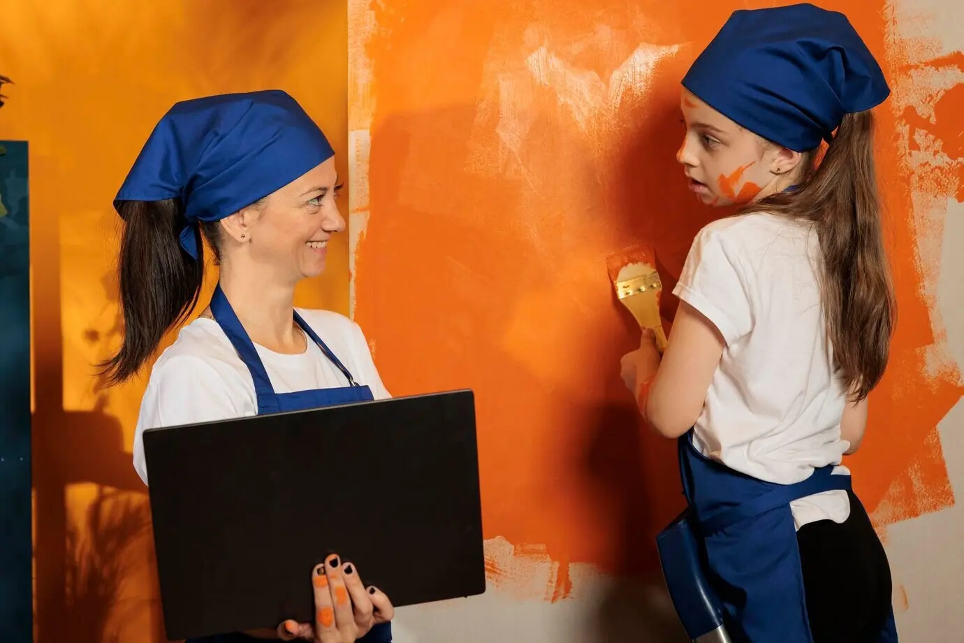 Mutter und Mädchen streichen orangefarbene Wände und nutzen einen Laptop, um online Deko-Inspiration zu finden. Das kleine Mädchen und die Frau suchen nach einem Renovierungsmodell mit Farbe und DIY-Werkzeugen für Hausarbeiten.